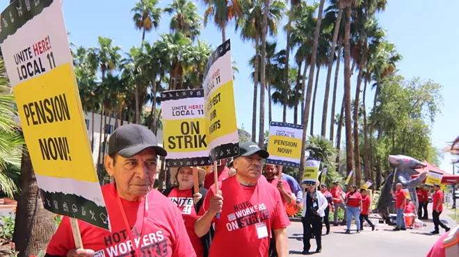 Hotel workers in red shirts hold picket signs with phrases like "On Strike!" and "Pension Now!" In the background is an inflatable rat.